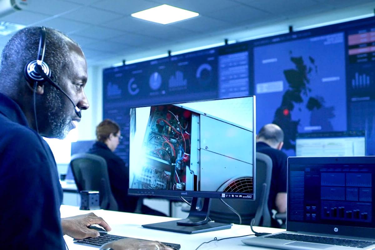 An engineer wearing a headset at a computer terminal, watching maintenance taking place elsewhere. A wall of computer displays and other people at desks are in the background